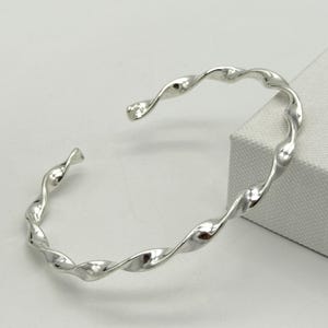 Puede incluir: Un brazalete de plata trenzado. El brazalete está hecho de una sola pieza de metal que se tuerce en forma de espiral.