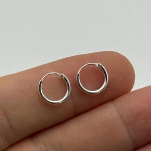 May include: A pair of small, circular silver hoop earrings. Each earring features a thin, polished metallic band that forms a complete, continuous circle. The design is simple and lightweight, showcasing a bright, reflective silver finish.