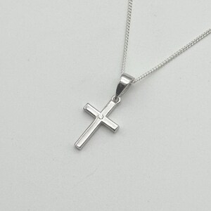 May include: A silver cross pendant necklace. The cross has a small, clear gemstone at the center. The chain is a delicate, silver curb chain. The pendant and chain are displayed against a light background.