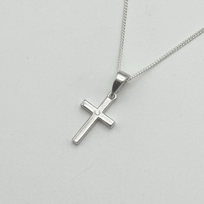 May include: A silver cross pendant necklace. The cross has a small, clear gemstone at the center. The chain is a delicate, silver curb chain. The pendant and chain are displayed against a light background.