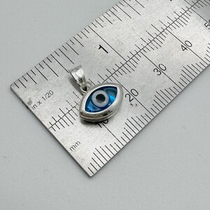 Puede incluir: Colgante plateado en forma de ojo, con dise&ntilde;o azul y blanco. El colgante est&aacute; sobre una regla, mostrando medidas en cent&iacute;metros. S&iacute;mbolo de protecci&oacute;n popular.