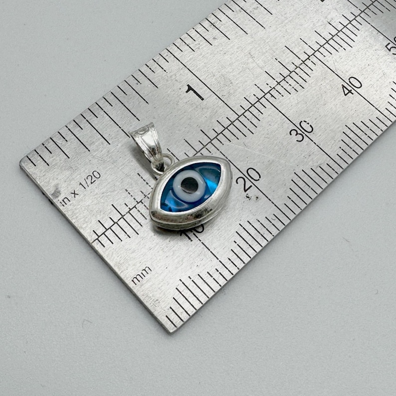 Puede incluir: Colgante plateado en forma de ojo, con dise&ntilde;o azul y blanco. El colgante est&aacute; sobre una regla, mostrando medidas en cent&iacute;metros. S&iacute;mbolo de protecci&oacute;n popular.