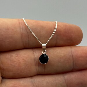 Peut inclure: Un collier en argent avec un petit pendentif rond en pierre noire. Le pendentif est facetté, captant la lumière. La chaîne est fine et argentée. Le collier est présenté sur un fond neutre.
