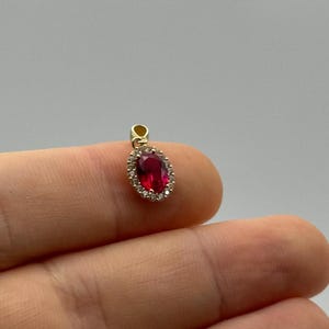 Op de afbeelding: Een gouden hanger met een ovale, dieprode edelsteen omringd door kleine, heldere edelstenen. De hanger heeft een kleine lus aan de bovenkant voor een ketting. De sieraden zijn geplaatst tegen een neutrale achtergrond.