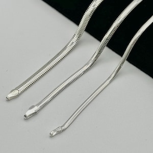 Puede incluir: Tres collares de cadena de serpiente plateados de diferentes longitudes. Cada cadena tiene un diseño plano en forma de escamas y un cierre de cabeza de serpiente. Los collares se muestran sobre un fondo blanco y negro, resaltando su brillo metálico.
