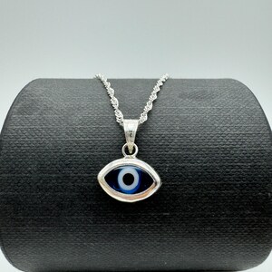 Puede incluir: Un collar plateado con un colgante de ojo turco. El colgante presenta un ojo azul y blanco enmarcado en plata. La cadena es de estilo cuerda trenzada y delicada. El collar se exhibe sobre una superficie negra.