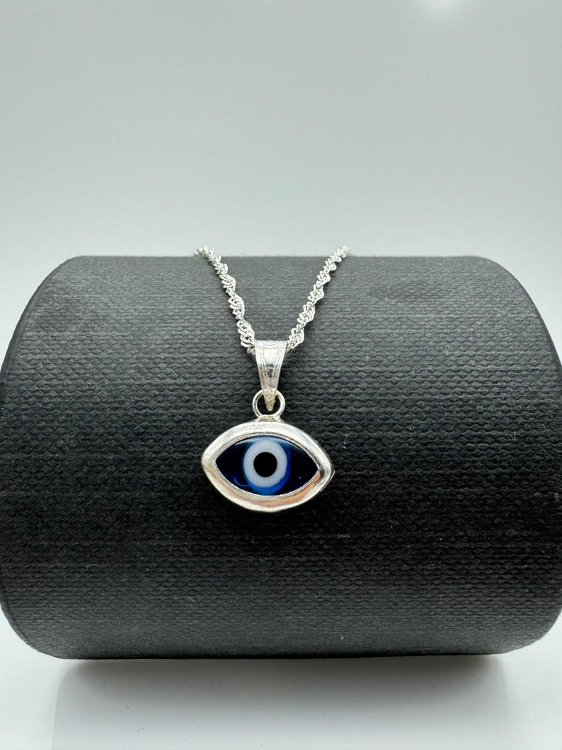Puede incluir: Un collar plateado con un colgante de ojo turco. El colgante presenta un ojo azul y blanco enmarcado en plata. La cadena es de estilo cuerda trenzada y delicada. El collar se exhibe sobre una superficie negra.