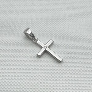 May include: A silver-colored cross pendant with a small, clear gemstone at the center. The pendant has a simple, classic design with a loop at the top for a chain. The cross is set against a textured, off-white background.