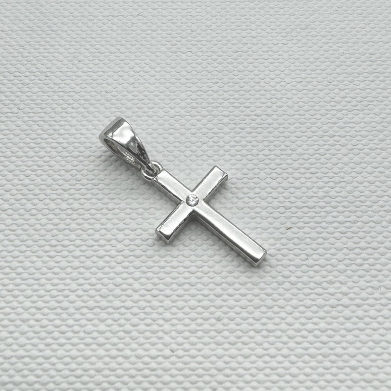 May include: A silver-colored cross pendant with a small, clear gemstone at the center. The pendant has a simple, classic design with a loop at the top for a chain. The cross is set against a textured, off-white background.