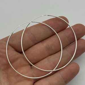 Peut inclure: Une paire de grandes créoles circulaires et fines, fabriquées en métal argenté poli. Ces boucles d'oreilles présentent un design simple en fil continu avec une surface lisse et réfléchissante, offrant une structure légère et épurée.