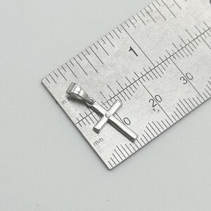May include: A small, silver-colored cross pendant with a single, small gem at the center. The cross is positioned on a metal ruler, showing measurements in inches and millimeters. The pendant is approximately 0.75 inches in length.
