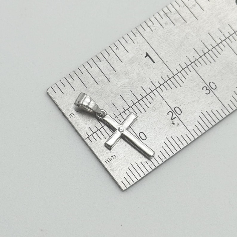 May include: A small, silver-colored cross pendant with a single, small gem at the center. The cross is positioned on a metal ruler, showing measurements in inches and millimeters. The pendant is approximately 0.75 inches in length.