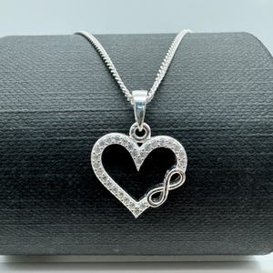May include: A silver necklace with a heart-shaped pendant. The pendant features a heart outline encrusted with clear gemstones and an infinity symbol. The necklace chain is a delicate, textured silver. The pendant is approximately 2.5 cm in width.