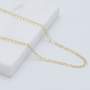 May include: A gold chain necklace with a curb link style. The chain is laying on a white surface.