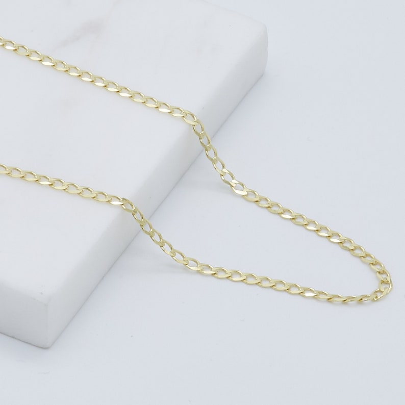 May include: A gold chain necklace with a curb link style. The chain is laying on a white surface.
