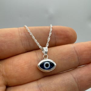 Puede incluir: Collar plateado con un colgante en forma de ojo. El colgante tiene un fondo azul con un iris blanco y negro. La cadena tiene un dise&ntilde;o delicado y retorcido. Una joya.