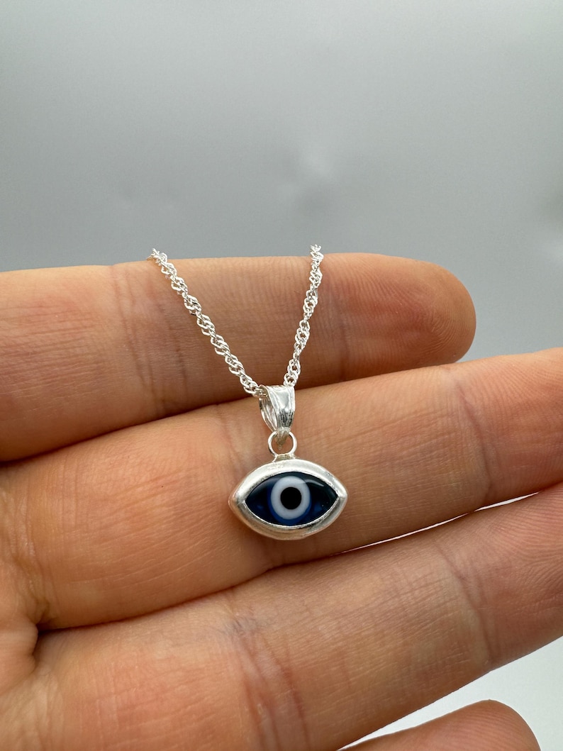Puede incluir: Collar plateado con un colgante en forma de ojo. El colgante tiene un fondo azul con un iris blanco y negro. La cadena tiene un dise&ntilde;o delicado y retorcido. Una joya.
