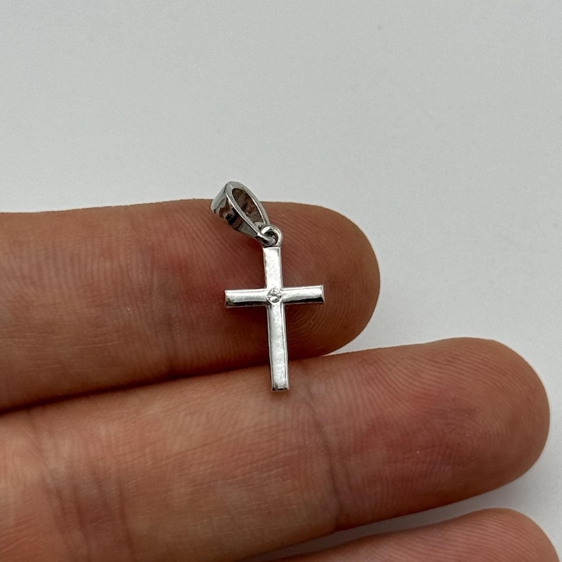 May include: A small, silver-colored cross pendant with a single, clear gemstone at the center. The cross has a simple, classic design and is suspended from a small loop. The pendant is likely made of metal and is shown against a neutral background.