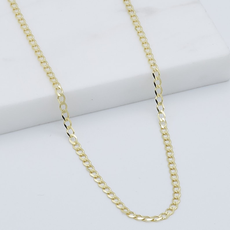 May include: A gold chain necklace with a curb link style. The chain is on a white surface.