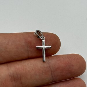 May include: A small, silver-colored cross pendant with a loop at the top for a chain. The cross has a simple, classic design with a small, round accent in the center. The pendant is shown against a neutral background.