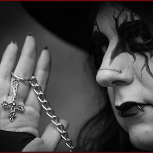 May include: A person with dark makeup and a nose ring wears a silver chain with a silver upside down cross pendant.