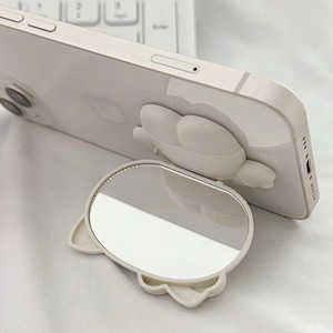 May include: A white phone stand with a mirror attached. The stand is shaped like a cartoon animal with a round mirror in the centre. The phone stand is designed to prop up a phone for hands-free viewing.