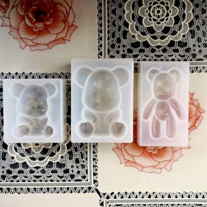 May include: Three white silicone moulds for making bear-shaped treats. The moulds are on a white surface with a lace pattern.