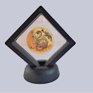 May include: A gold-coloured coin featuring a colourful image of a snake and Chinese characters is presented in a black diamond-shaped frame with a black base.