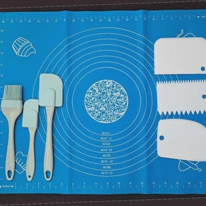 May include: A blue silicone baking mat with metric measurements and a set of light blue baking tools. The tools include a brush, two spatulas, and three scrapers. The mat has a cupcake illustration and a decorative circular design.