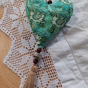May include: A heart-shaped fabric decoration with a teal and white floral design. It features a jute tassel and wooden beads. The ornament is suspended against a white surface with a lace trim.
