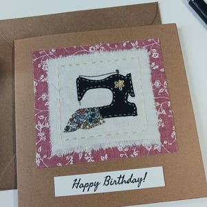 May include: A birthday card made from brown kraft paper, featuring a fabric square with a black sewing machine design. The fabric is bordered by a pink floral print. The card includes the text "Happy Birthday!" in white.