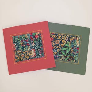 May include: Two decorative cards with fabric squares. One card is red, the other is green. Each card features a square of patterned fabric with floral and leaf designs in blue, red, and yellow. The fabric squares are stitched onto the cards with gold thread.