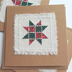 May include: A pile of square, brown cards, each with a white fabric patch. The patch features a star design made from red and green patterned fabric, stitched onto the white fabric. The edges of the fabric are frayed.