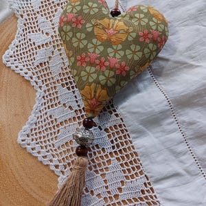 May include: A heart-shaped decoration with a floral pattern in green, yellow, and pink. It features a jute string, wooden beads, a silver bead, and a brown tassel. The ornament is displayed on white crocheted lace and a light-colored surface.