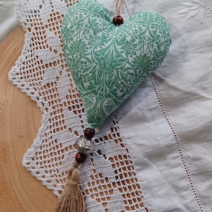 May include: A heart-shaped fabric decoration with a green floral design. It features a jute string, wooden beads, a silver bead, and a tassel. The ornament is presented on a white lace doily and a wooden surface.