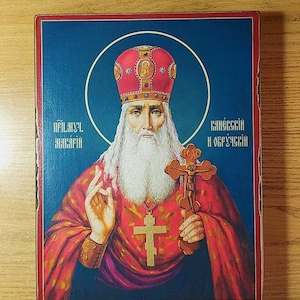 Może przedstawiać: Ikona religijna przedstawiająca brodatego mężczyznę z białą brodą i czerwoną koroną zdobioną złotem. W prawej ręce trzyma krzyż, a lewą podnosi w geście błogosławieństwa. Ikona jest namalowana na drewnianym panelu i ma niebieski tło. Tekst na ikonie brzmi "Преп. Макарий Калязинский и Овручский".