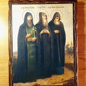 May include: A religious icon depicting three monks in black robes with gold accents. The icon is painted on a wooden panel with a brown frame. The text on the icon reads "св. прп. Алексий Человек Божий, св. прп. Тихон, св. прп. Марк Пекоша.