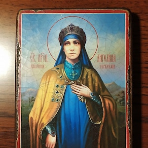 May include: A religious icon depicting a woman in a blue robe and yellow cloak, wearing a crown and holding her hand to her chest. The icon is framed in a wooden frame with a red border. The text on the icon reads "Св. Прп. Княгиня Ангелина Сербская".