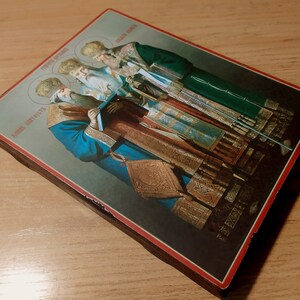 Puede incluir: Un icono religioso vintage que representa a cuatro figuras con ropas adornadas. El icono est&aacute; pintado en un panel de madera con un borde rojo y presenta el texto "Церковь Святых" y "Иоанн Златоуст" en escritura cir&iacute;lica.