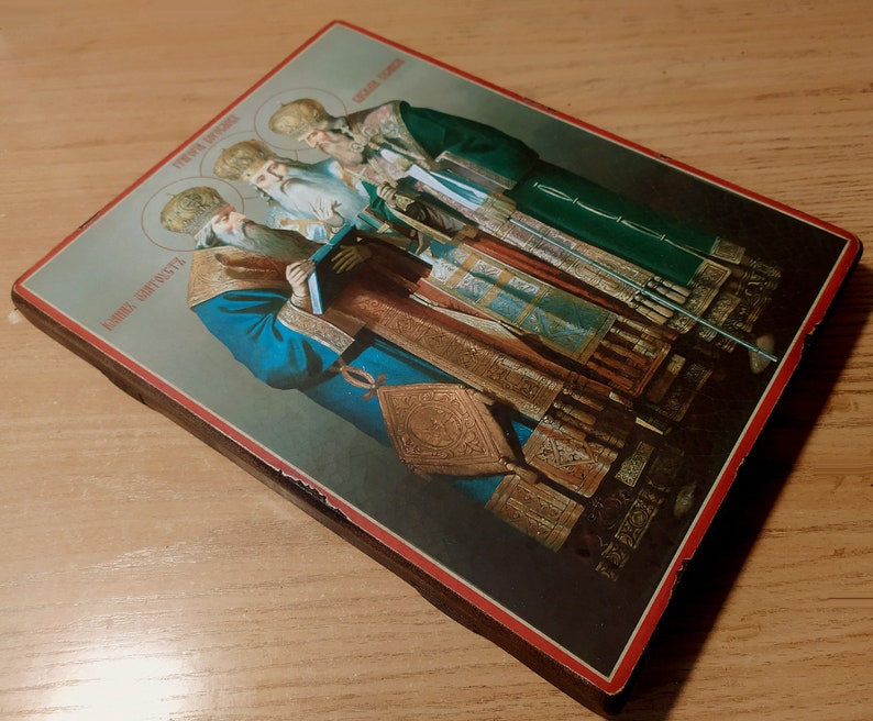 Puede incluir: Un icono religioso vintage que representa a cuatro figuras con ropas adornadas. El icono est&aacute; pintado en un panel de madera con un borde rojo y presenta el texto "Церковь Святых" y "Иоанн Златоуст" en escritura cir&iacute;lica.