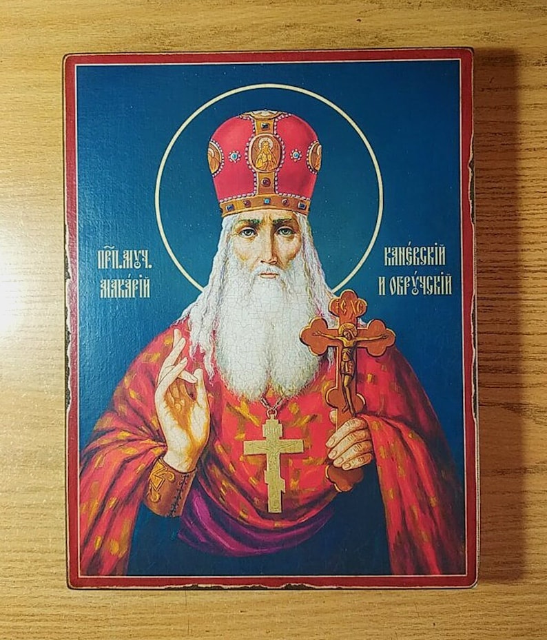 Może przedstawiać: Ikona religijna przedstawiająca brodatego mężczyznę z białą brodą i czerwoną koroną zdobioną złotem. W prawej ręce trzyma krzyż, a lewą podnosi w geście błogosławieństwa. Ikona jest namalowana na drewnianym panelu i ma niebieski tło. Tekst na ikonie brzmi "Преп. Макарий Калязинский и Овручский".