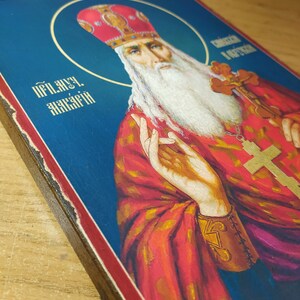 Może przedstawiać: Ikona religijna przedstawiająca brodatego mężczyznę z białą brodą i czerwoną koroną zdobioną złotem. Mężczyzna jest ubrany w czerwoną szatę z złotymi zdobieniami i złotym krzyżem. Tło jest niebieskie. Tekst "Преп. Акакий" jest napisany w lewym g&oacute;rnym rogu obrazu.