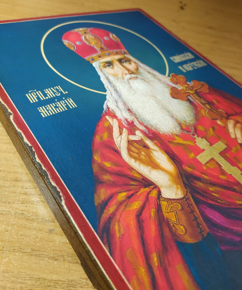 Może przedstawiać: Ikona religijna przedstawiająca brodatego mężczyznę z białą brodą i czerwoną koroną zdobioną złotem. Mężczyzna jest ubrany w czerwoną szatę z złotymi zdobieniami i złotym krzyżem. Tło jest niebieskie. Tekst "Преп. Акакий" jest napisany w lewym g&oacute;rnym rogu obrazu.