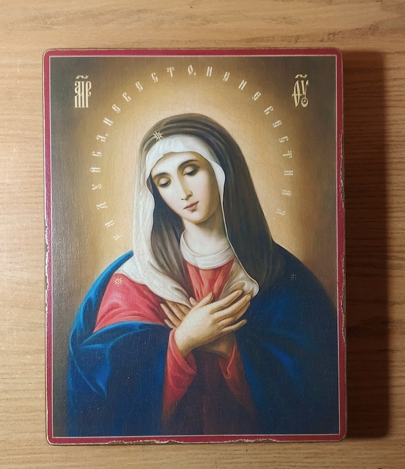 May include: A religious icon depicting the Virgin Mary with a blue robe and a white veil. She is holding her hands together in prayer. The icon is framed in a red wooden frame and has a gold background.