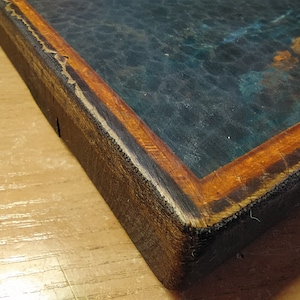 May include: Close-up of the corner of a wooden frame with a dark blue and green painting inside. The frame is made of dark wood with a thin orange border.