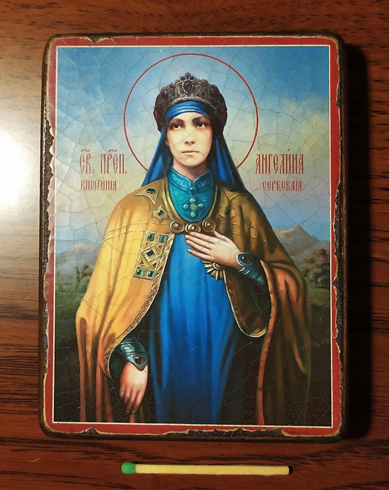 May include: A religious icon depicting a woman in a blue robe and yellow cloak, wearing a crown and holding her hand to her chest. The icon is framed in a wooden frame with a red border. The text on the icon reads "Св. Прп. Княгиня Ангелина Сербская".