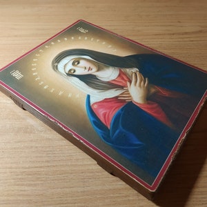 May include: A wooden framed icon depicting the Virgin Mary with a blue robe and red undergarment. The icon has a gold halo around the Virgin Mary's head and a gold background. The icon is surrounded by a border of Cyrillic letters.