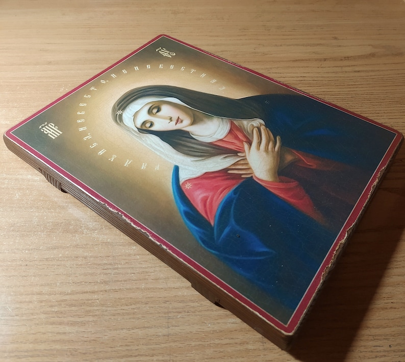 May include: A wooden framed icon depicting the Virgin Mary with a blue robe and red undergarment. The icon has a gold halo around the Virgin Mary's head and a gold background. The icon is surrounded by a border of Cyrillic letters.
