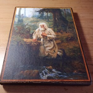 May include: A religious icon depicting a bearded man in a white robe kneeling in a forest setting. The icon is painted on a wooden panel and features a stream of water in the foreground. The text "Слава Господу Богу!" is written in Cyrillic script at the top of the icon.
