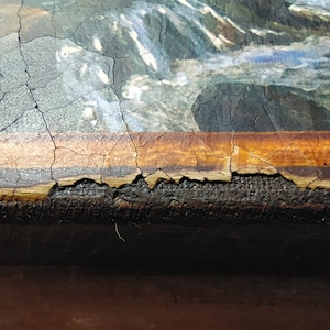 May include: Close-up of a painting with a cracked, brown frame. The painting depicts a landscape with water and rocks. The frame is damaged and peeling.